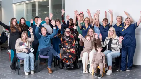 Children of the 90s A group of researchers, midwives and participants of the study, all sitting together and raising their hands in the air, smiling to camera