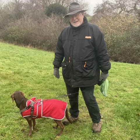 Steve Moody is standing on grass with his dog poppy in front of him who is on a lead and wearing a red coat
