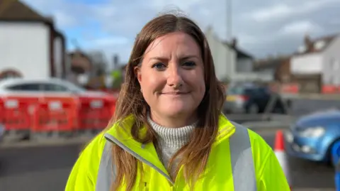 George Carden/BBC Andrea smiling at the camera wearing a high-vis with traffic behind her. She has medium length brown hair and a turtle neck jumper