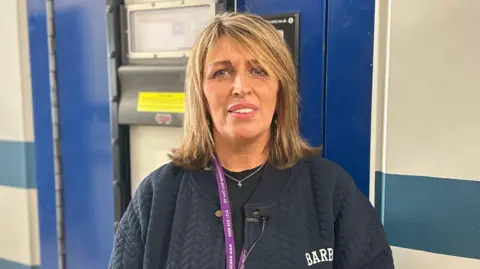 Woman has medium length blonde and brown highlighted hair. She is smiling with pink lipstick and showing her teeth. She is wearing a navy blue jacket and a thin silver necklace around her neck. Standing in front of a police custody door.