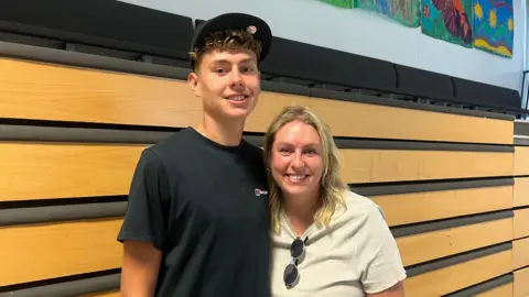 A teenager in a black t-shirt and black cap smiles for the camera with his mum.