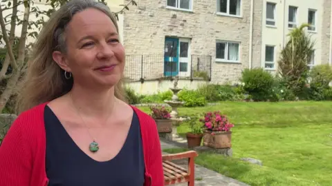 The picture shows an adult standing in a landscaped residential garden, wearing a dark top with a red cardigan and a round pendant necklace. Behind the person are a green lawn, flower planters, a small stone fountain, a wooden bench, and a light-coloured apartment building with multiple windows and a blue door.
