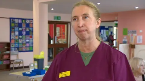 A woman with long blonde hair wearing purple scrubs over a green shirt with a yellow name tag that says "Jane Berry".