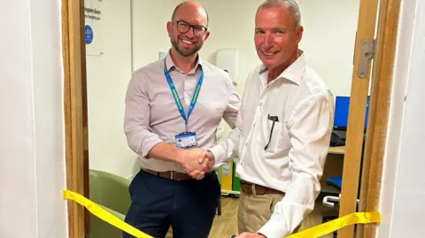 A man wearing a shirt and a blue lanyard shakes the hand of a man wearing a white shirt. The pair are standing in the new suite, with a yellow ribbon seen in front of them.