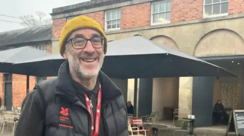 A man in a black coat and yellow woollen hat with a grey beard, in front of a large building with two black canopies outside and chairs below it
