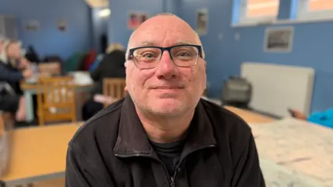 BBC/Jim Scott David Cope, who is an Army veteran, is wearing black half framed glasses and a dark brown zipped fleece. In the background are veterans who are sitting at a table behind Mr Cope and talking. 
