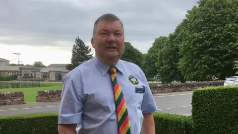 Fellrunner Village Bus Kevin McGilloway. He has short grey hair and is wearing a blue short-sleeved shirt with a red, green and yellow striped tie. He is standing in a garden bordered by neat hedges.