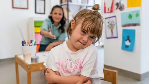 Young girl is close to camera with arms folded, adult is in background sitting at a low table with children's art on the walls