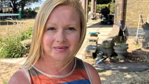 John Devine/BBC Jo Croft has shoulder length blonde hair and a multi-coloured sleeveless dress. She is standing in a garden which has several bushes and hedges.