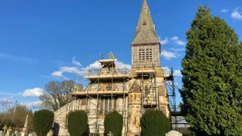 Tim Bridges The outside of a church which has scaffolding on around it. The church has a steeple and has cream and yellow stone work. It has a large fir tree at the front of the picture and four others in front of the church. 