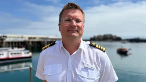 A grey-haired man in glasses, in a white naval shirt with stripes on his shoulders. He is standing in front of St Peter Port harbour and Castle Cornet.