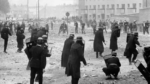 A black-and-white photo showing a number of police officers with helmets, shields and batons are facing towards a number of people throwing items in their direction. Debris is visible on the ground, and a number of houses can also be seen in the background.