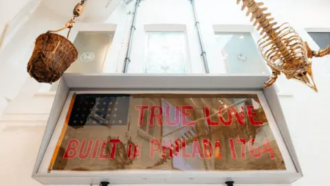 Tom Arran Photography Writing on top of the flag says "Trud Love - built in PhiladA 1764. It hangs on a white wall below three internal windows.