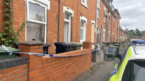 Blue and white police tape is draped along a brick wall outside a row of terraced houses. A white, yellow and blue police car is parked on the road outside the house.