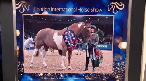 A picture on a mantlepiece which reads LONDON INTERNATIONAL HORSE SHOW RANCHELLA ROLEX CHAMPION 2025
