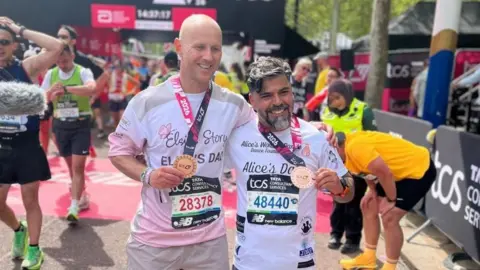 A bald man with a T-shirt saying Elsie's dad - holds his medal and his arm around his friend who has dark and grey hair and has a T-shirt which reads Alice's dad 