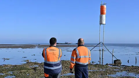 Two men wearing hi-vis orange jackets have their backs to the camera. They look out from a beach to the sea - where there are sand banks and a ship to be used for target practice in the background.