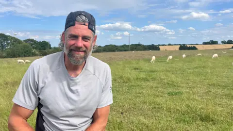 Katie Radley/BBC A man wearing a grey T-shirt is smiling at the camera. He is wearing a dark grey cap on backwards. Behind him is a field with sheep in it. In the background are trees, more fields and a telegraph pole.