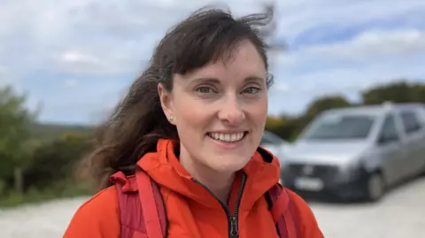 Woman with long brown hair, tied up at the back, is smiling at the camera. She is wearing an orange waterproof and a rucksack 