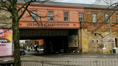 Psa A railway station, with GILMOUR STREET STATION in large letters across the building. A graffiti mural next to it has eyes and a smiling face, looking like a Thomas the Tank Engine character. 