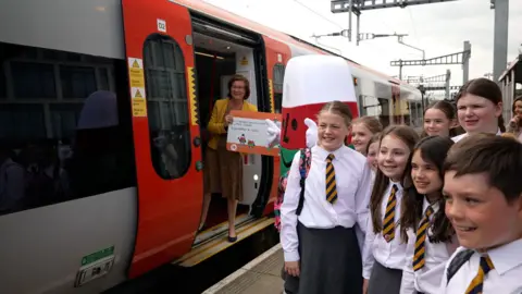 Eight pupils in white shirts and yellow and black ties stood on the platform. Eluned Morgan is inside the train holding up a large train ticket with Mistar Urdd standing next to her.