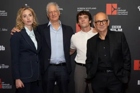 EIFF/Cameron Allan Freya Mavor is in a blue suit and white shirt and has long blonde hair. Marc Turtletaub has white hair and is in a blue blazer, grey trousers and light blue open-necked shirt. Fionn Whitehead sports a white short-sleeved shirt buttoned at the neck and Luis Gnecco is in a black suit and shirt. They stand in a row, arms around one another, in front of a black, white and red Edinburgh International Film Festival board. 