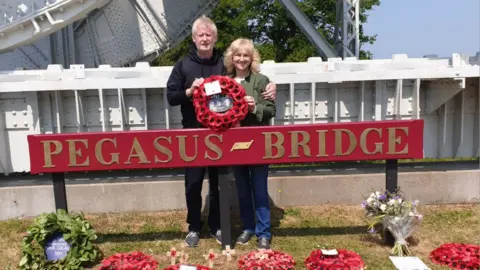 Carole Stewart Carole and Gerard Stewart a couple in their 60s are standing in front of a huge sign which says Pegasus Bridge holding a wreath of poppies. A number of other wreaths are at their feet. The metal bars of the bridge are behind them 