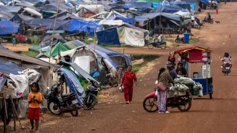 A dirt road cutting through the site of the camp for the displaced families, made of blue tarpaulin makeshift tents on sticks. A woman is walking down the road, while another woman is buying vegetables from a man on a motorcycle selling groceries in plastic bags. A little girl can be seen in the foreground walking. 