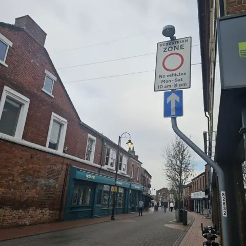 A sign above a cobbled street states that no vehicles are allowed on the road between the hours of 1000 and 1600.