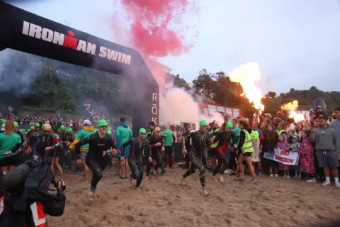 Huw Fairclough A group of people wearing wetsuits and bright green swimming caps run across some sand in front of crowds of people. A black archway reads "IRONMAN SWIM" in large lettering. Red smoke and what looks like fire are rising into the air.