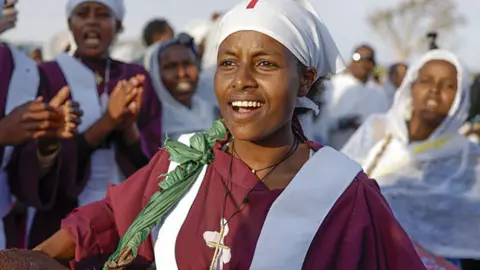 A woman in a purple top chants an bangs a drum.