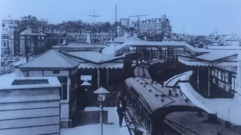  A black and white postcard image of an old train station, with a train stopped at one of the platforms. 