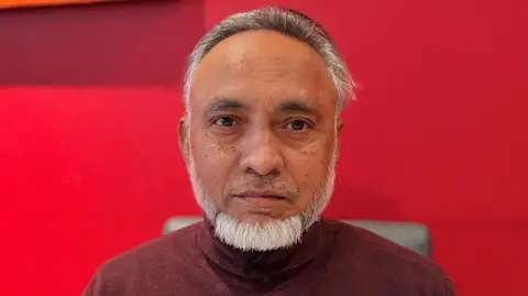 A man with white hair and a white beard standing in front of a red background.