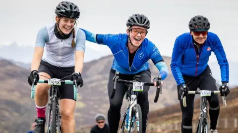 Three riders cycling close together. One of the riders has an arm round a fellow cyclist as they cycle towards the photographer taking the image. Behind the group are brown hills.
