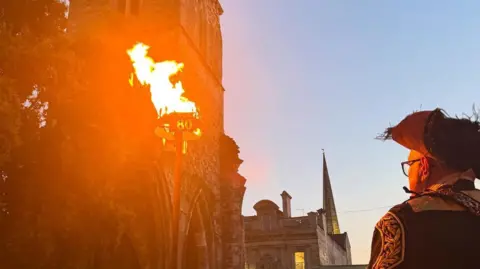 A burning beacon. In the foreground is a man in a tricorn hat.