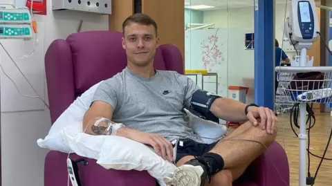 Liam Landers smiling at the camera while sitting in a purple chair on a hospital ward. He has tubes going into his arms, as he has chemotherapy at hospital. 