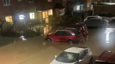 A high shot of a street being flooded by muddy water. Several cars have the water up to the middle of their wheels and the water can be seen up against the bottom of several front doors