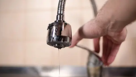 A tap with very little water coming out of it. A person is pushing the tap up.