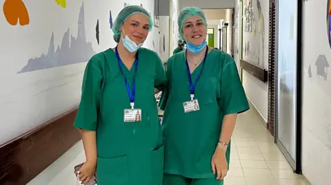 Two women wearing green scrubs with face masks under their chins pictured in a hospital corridor