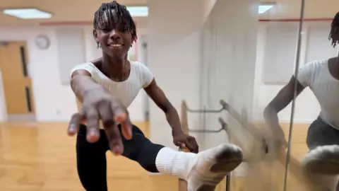Jaydon, a fifteen-year-old black ballet dancer, stretches at a studio barre, extending one leg onto the rail and reaching forward with an outstretched arm, while standing on a wooden floor beside a mirrored wall. He is wearing peach-coloured ballet shoes which are a bit grubby on the bottom, black leggings and a skin-tight white top.