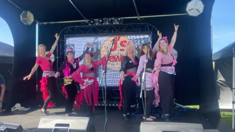 Sara Dunleavy Ladies wearing pink outfits dancing on a stage. They all have one hand in the air as they perform.