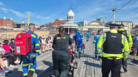 HM Coastguard A member of ambulance staff pushes a person on a stretcher with police officers and coastguard officers walking alongside side her. They are taken from behind. They are walking up Blackpool's North Pier on a sunny day. Visitors line the pier and the seafront.