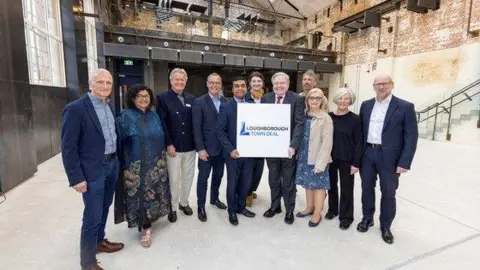 supplied 11 people posing to the camera two in the middle holding a panel saying "Loughborough Town Deal"