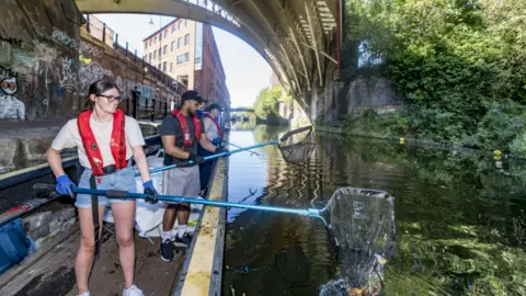 Canal & River Trust A woman with a large net picking litter up from a canal from a boat, with two other people next to her