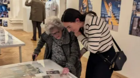 Hannah Kent Two people lean over a glass display case at Bury FC looking at club photos and printed memorabilia. One is Joan who wears a gray textured coat, the other is Hannah wearing a a striped black-and-white top with a crossbody bag. The room has parquet flooring and walls covered with large photo exhibits.