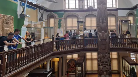 Museum of Archaeology and Anthropology A number of people in the Museum of Archaeology and Anthropology, which shows a wooden mezzanine floor, with displays and a wooden pole in the middle.