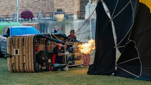 PA Media A man and a woman tending to a hot air balloon. The man is sat by the basket, which is on its side, as flames shoot out of the burner into an inflating balloon canopy. The woman is behind stood behind the basket operating a large fan. 