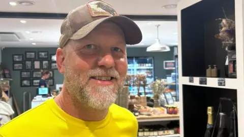 A man with a grey beard and blue eyes wearing a brown cap and yellow T-shirt smiles at the camera. He is standing in a farm shop with a till and produce behind him. 
