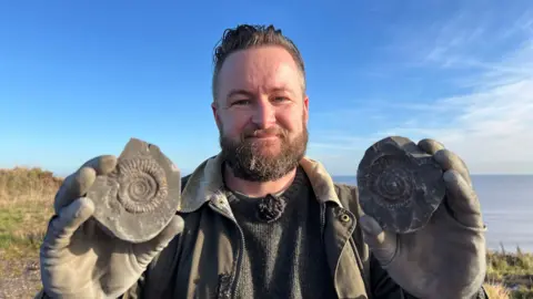A man with dark hair and beard smiles into the camera as he holds up two halves of a rock containing an ammonite fossil. The fossil is dark with a spiral pattern. The man is wearing a dark green knitted jumper and a similar coloured waxed jacket. He is standing on a grassed area next to the sea under a sunny blue sky.