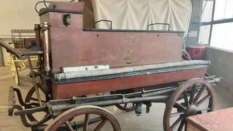 Katie Radley/BBC An old fashioned looking rustic red fire engine that was pulled by horses in the Victorian era. It has large wheels and a metal hose running down the side above the wheels.